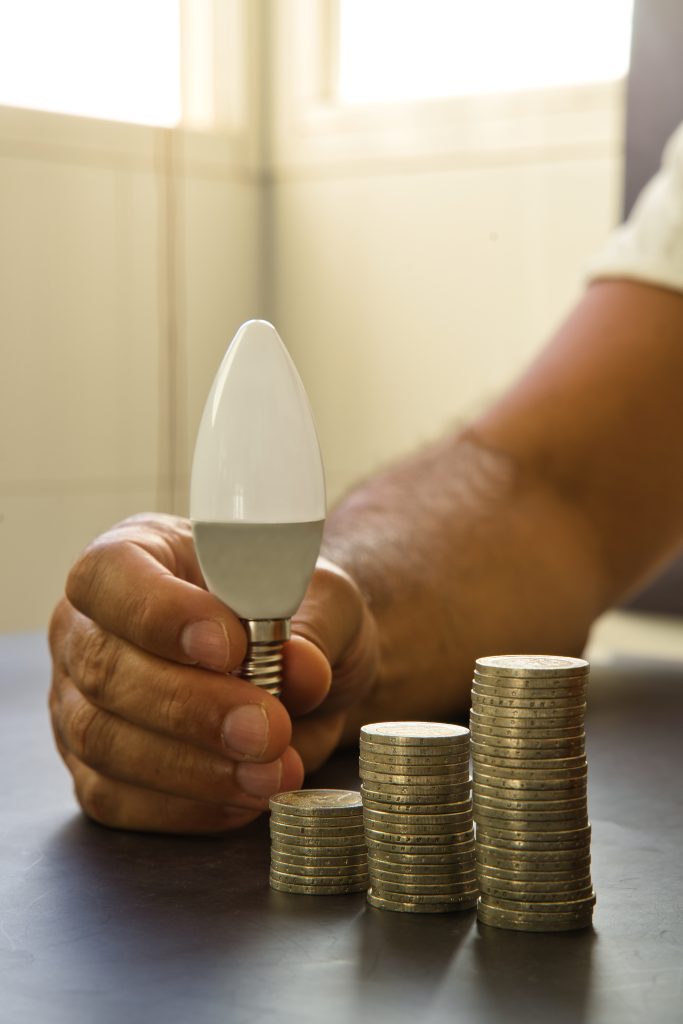 A man's hand holding a light bulb next to a stack of coins, as a concept of rising energy costs.